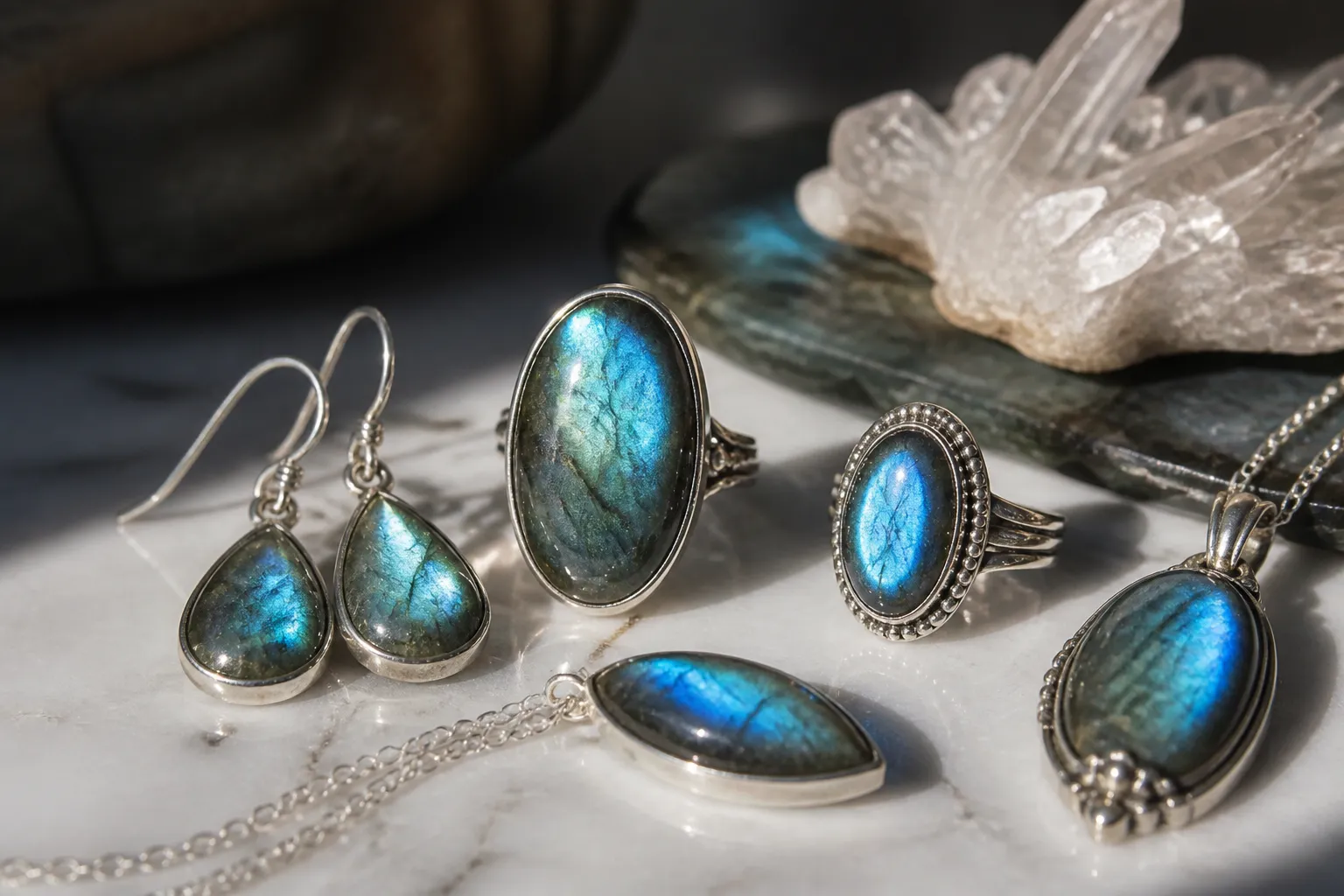labradorita piedra - Foto editorial de una colección de joyería con labradorita piedra natural —pendientes drop, anillos y colgantes— sobre superficie de mármol blanco con iluminación lateral que resalta los destellos iridiscentes