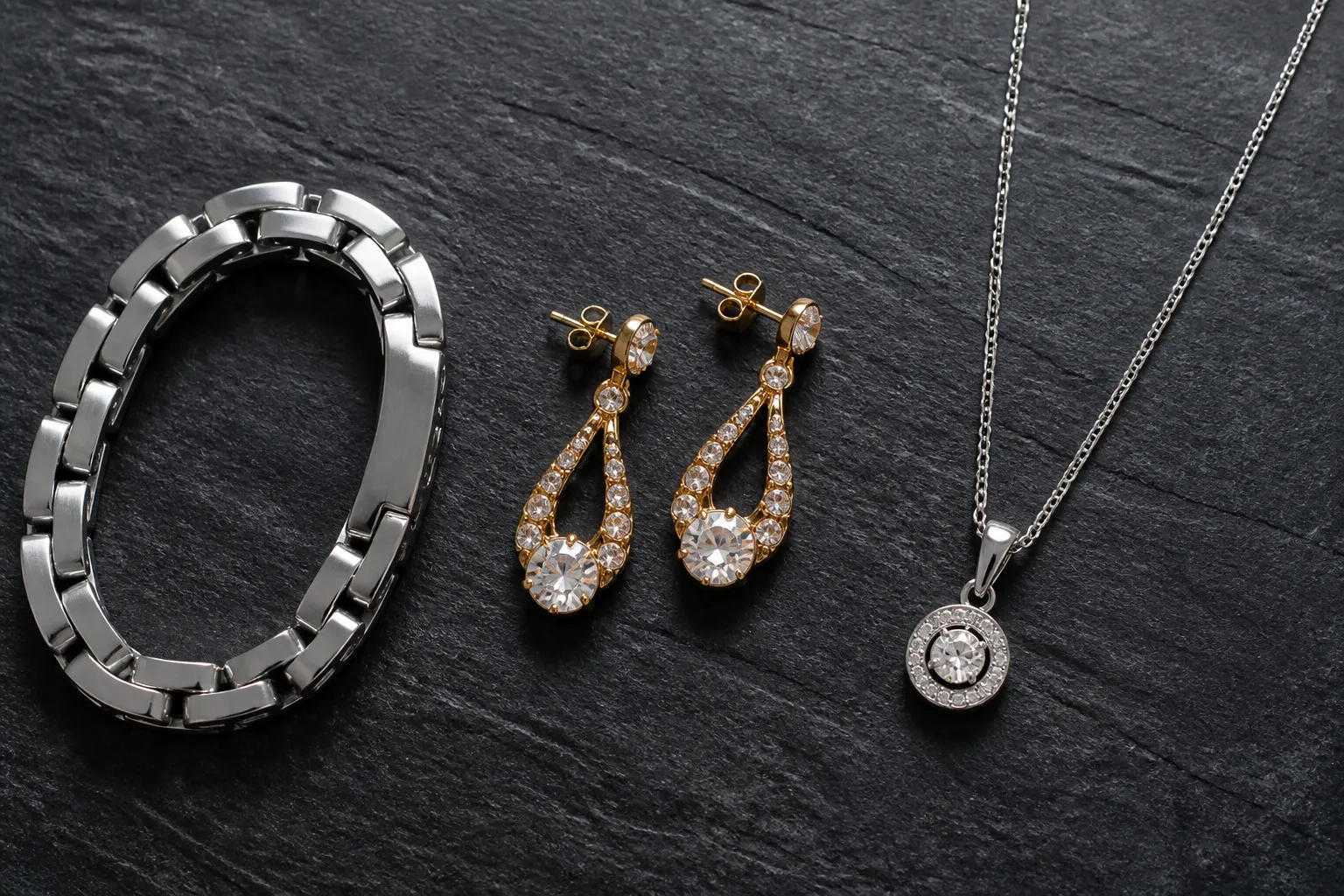 bijoux acier inoxydable - Flat lay photographique de trois familles de bijoux disposées côte à côte — bracelet en acier inoxydable poli, boucles d'oreilles pendantes en cuivre plaqué or avec détails cristal, collier en argent 925 — sur fond ardoise avec étiquettes de matière visibles