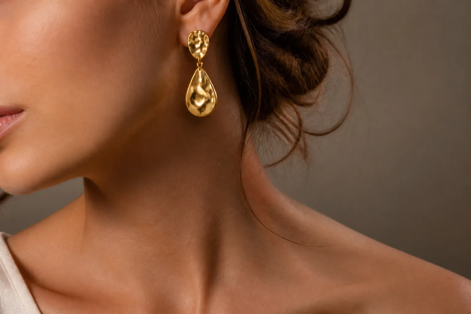 joyas de oro - Fotografía de detalle de joyería de moda — pendientes dorados tipo drop sobre cuello de mujer, con fondo neutro claro, mostrando la textura y el brillo del acabado chapado en oro