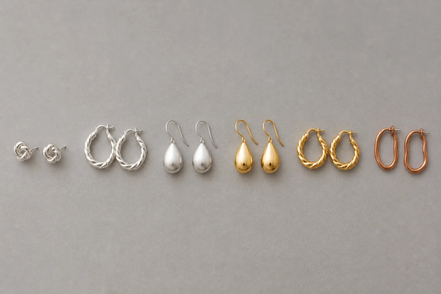 was ist sterling silber - Flatlay-Foto eines Sortimentsmusters: verschiedene Silber- und vergoldete Ohrringe nebeneinander auf neutralem Hintergrund, mit Materialetiketten versehen — zeigt die Bandbreite zwischen Sterling Silber und veredeltem Kupfer im direkten Vergleich