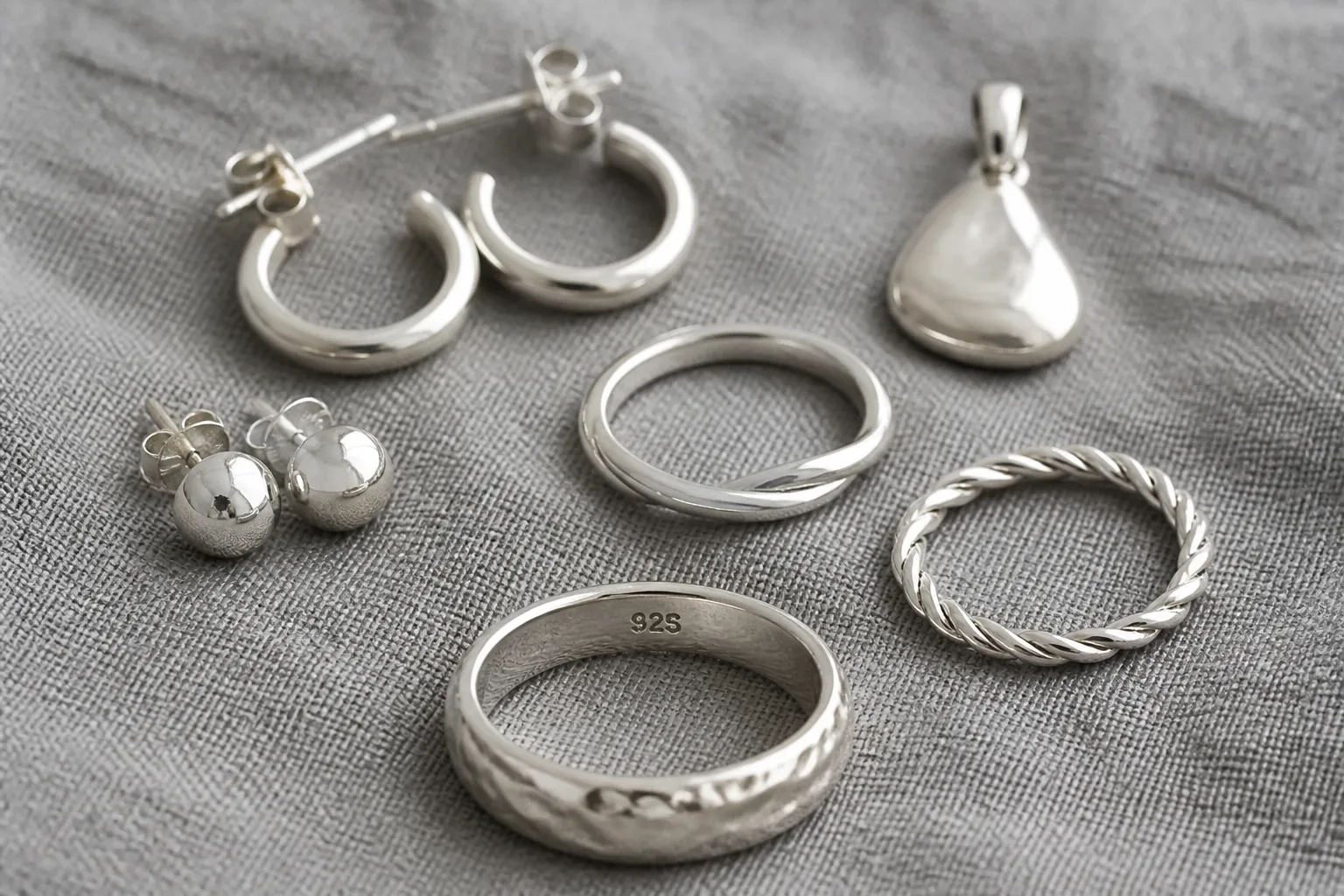 echt silber schmuck - Flatlay-Fotografie verschiedener echter Silberschmuckstücke — Ohrstecker, Creolen, Anhänger und Ringe auf strukturiertem grauem Untergrund, mit sichtbarem 925-Stempel an einem Ring im Vordergrund