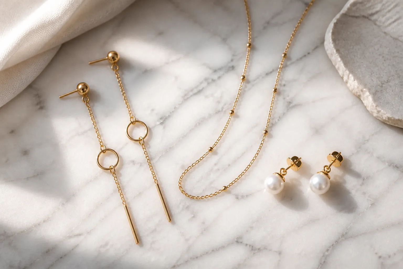 schmuck gold damen - Photorealistic flat-lay of assorted women's gold jewelry pieces — drop earrings, a delicate goldkette necklace, and pearl-drop stud earrings — arranged on white marble, natural daylight from the left side, no text or labels, editorial product photography composition