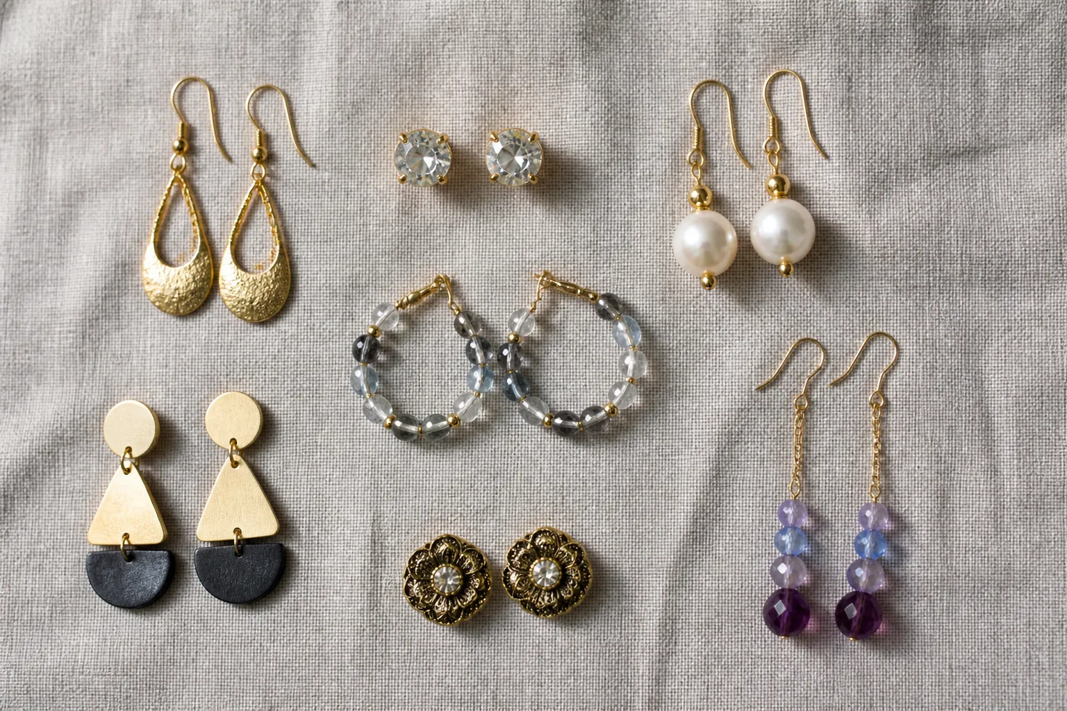 schmuck ohrringe - Photorealistic flat-lay photo of seven distinct earring styles arranged on a light grey linen surface — gold-plated drop earrings, rhinestone studs, freshwater pearl drops, glass bead hoops, two-tone geometric shapes, vintage flower frame studs, and gradient crystal dangles. Natural side lighting from the left, soft shadows, no text overlays, documentary photography style.