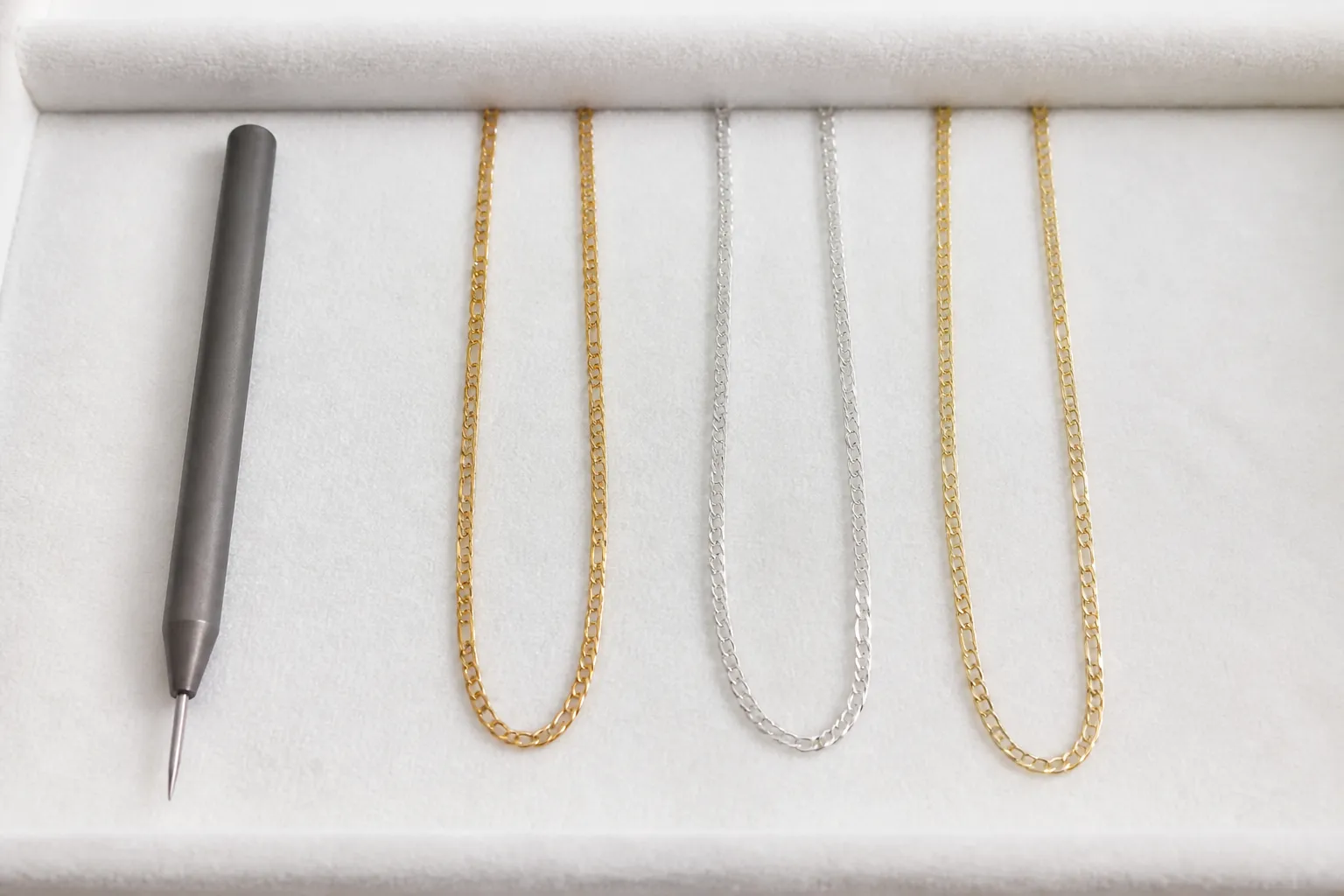 orion mpulse permanent jewelry welder - Overhead editorial photo of a flat-lay showing three types of fine jewelry chain (gold-filled, sterling silver, 14k solid gold) arranged side by side on a white velvet surface, with a tungsten electrode stylus placed beside them for scale, natural daylight, no text overlays
