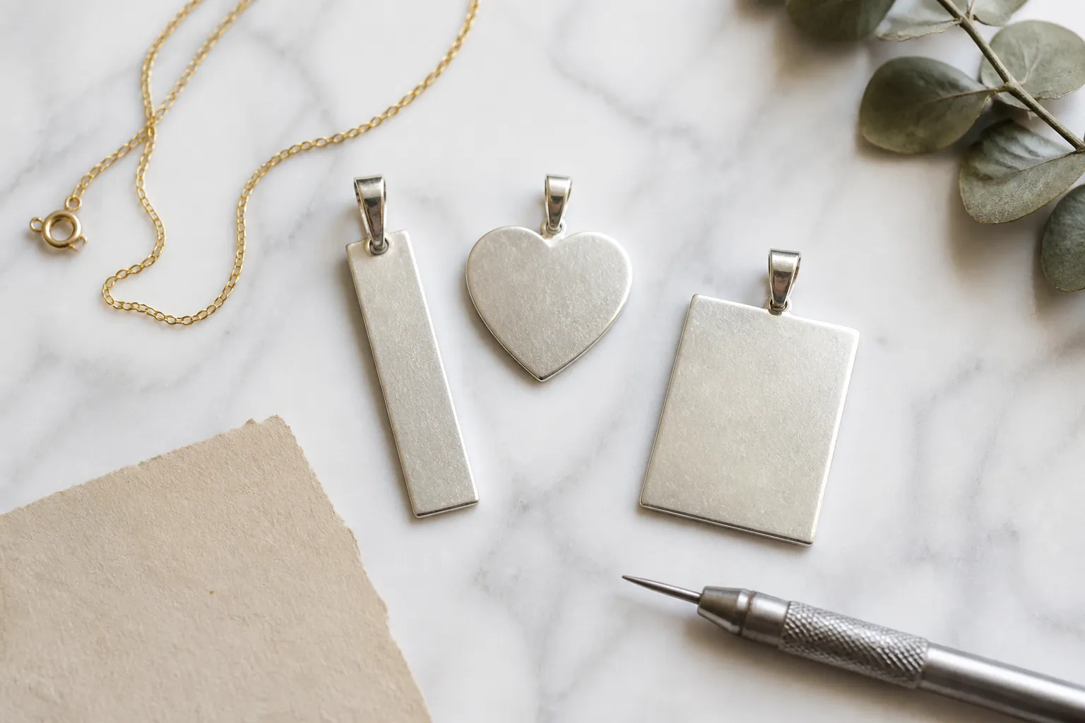 blank jewelry for engraving - Editorial flat-lay photograph: top-down view of white marble surface with three engraved sterling silver pendant blanks (a bar, a heart, and a rectangle) arranged alongside a fine gold chain, a small graver tool, and a handwritten note card; natural daylight from above, documentary styling, no overlaid text or labels