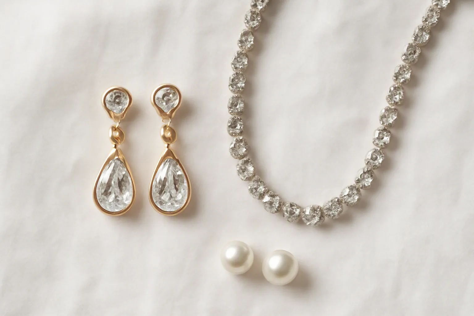 bijoux mariage - Editorial flat-lay photo on a white linen surface: three wedding jewelry sets arranged symmetrically — gold-plated drop earrings, a crystal rhinestone necklace, and pearl stud earrings — small discreet product tags visible, natural overhead daylight, clean and curated styling evoking a professional wholesale catalog
