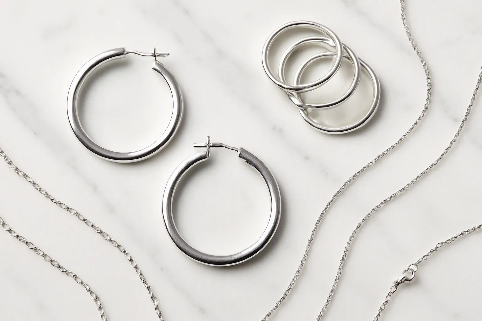 joyería de plata - Flat lay editorial de colección de joyería de plata sobre mármol blanco: pendientes de aro, anillos apilables y cadenas finas con iluminación de estudio, mostrando variación de acabados rodio y plata 925