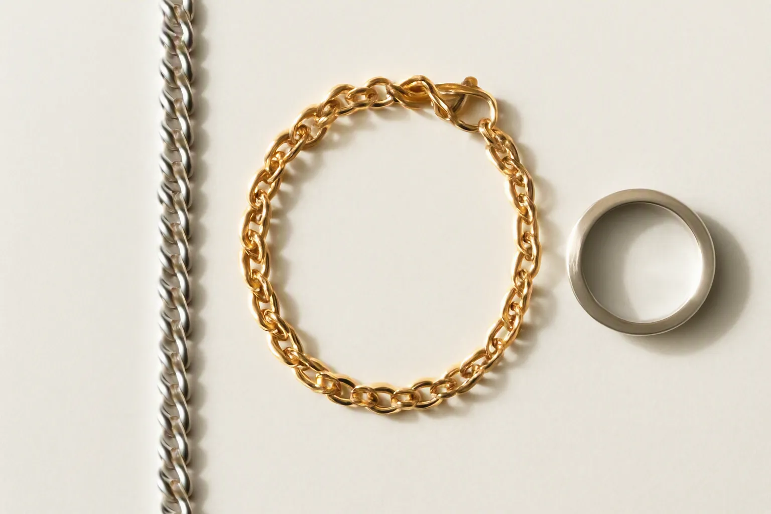 jewellery wholesale - Editorial top-down flat lay of three distinct jewellery material samples arranged side by side on a white surface — a polished sterling silver chain, a gold-filled bracelet with lobster clasp, and a stainless steel ring with brushed finish. Small material label cards beside each piece. Natural daylight, authentic product photography style.
