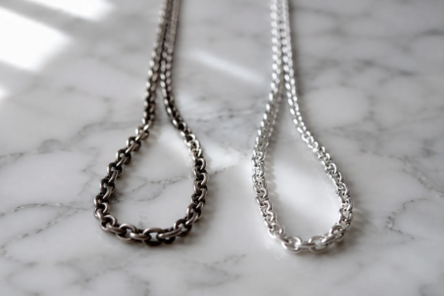nettoyer bijoux argent - Photorealistic close-up of two 925 silver chain necklaces on a white marble surface — one heavily oxidized and dark, the other freshly polished and gleaming — natural daylight from a window on the left, shallow depth of field, documentary jewelry care photography
