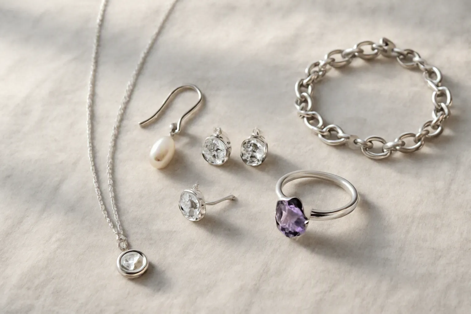 bijoux argent femme - Photorealistic flat-lay editorial photo of a curated women's silver jewelry collection arranged on pale gray linen fabric — a delicate chain necklace with a small pendant in the foreground, two pairs of silver stud earrings with faceted crystal stones in the center, a pair of longer drop earrings on the left, a silver link bracelet and a ring set with a deep purple amethyst on the right — soft diffused natural window light from the left, clean product styling with no shadows