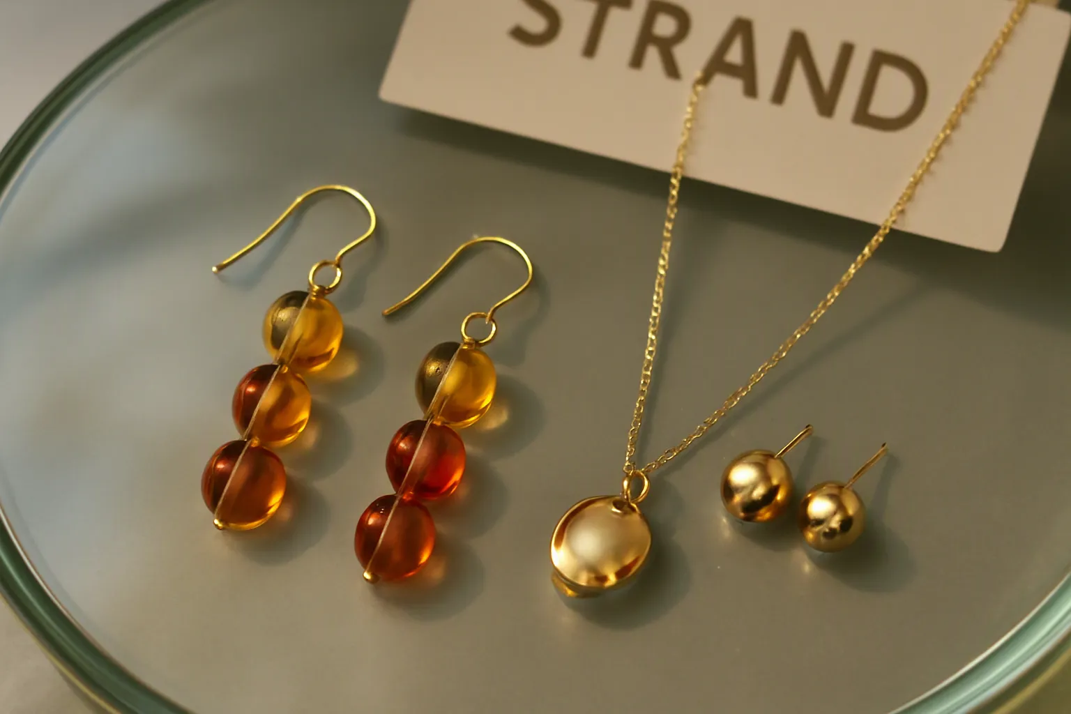 سوق الصين - صورة واقعية لعرض إكسسوارات STRAND على طاولة زجاجية — أقراط متدلية بالخرز الزجاجي بتدرجات ألوان خريفية، وطقم قلادة وأقراط بطلاء ذهبي على نحاس، والتسمية التجارية ظاهرة بشكل طبيعي في الخلفية، إضاءة استوديو ناعمة تعكس لمعة المعدن