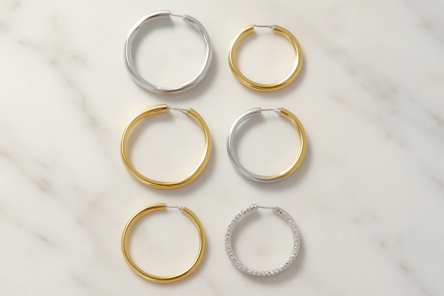 pendientes de aro - Fotografía editorial de una selección de pendientes de aro en distintos acabados —plata, dorados, bicolor y con cristal— sobre superficie de mármol blanco, mostrando la variedad de diámetros y materiales disponibles en catálogo