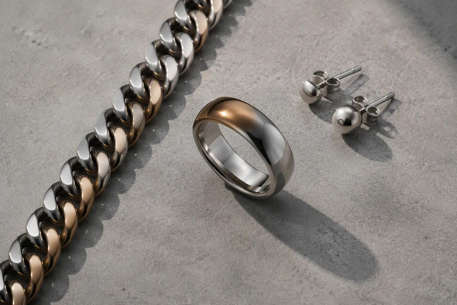 achat argent - Editorial flat-lay photo on brushed concrete surface: a wide two-tone rhodium-plated copper ring with gradient finish center-frame, a Cuban link chain necklace extending diagonally left, and a pair of minimalist stud earrings top-right, natural raking side light casting soft defined shadows, items arranged in a loose triangular composition, no branding or text visible, matte surface texture in foreground