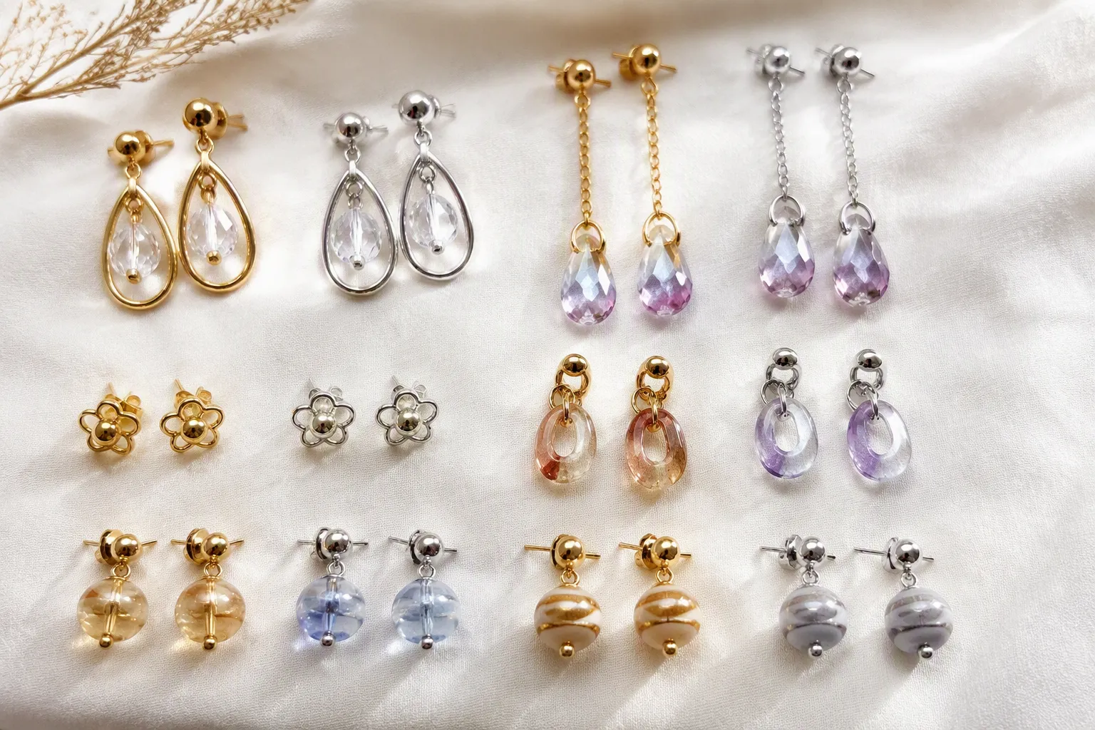 التسوق من الصين - Editorial flat-lay photo of an assortment of gold-plated and rhodium-plated copper earrings in different styles — drop earrings, stud earrings, gradient crystal and two-tone glass bead designs — arranged in an organized grid on a white linen surface, soft diffused natural light from the side, authentic product photography feel without text or price tags