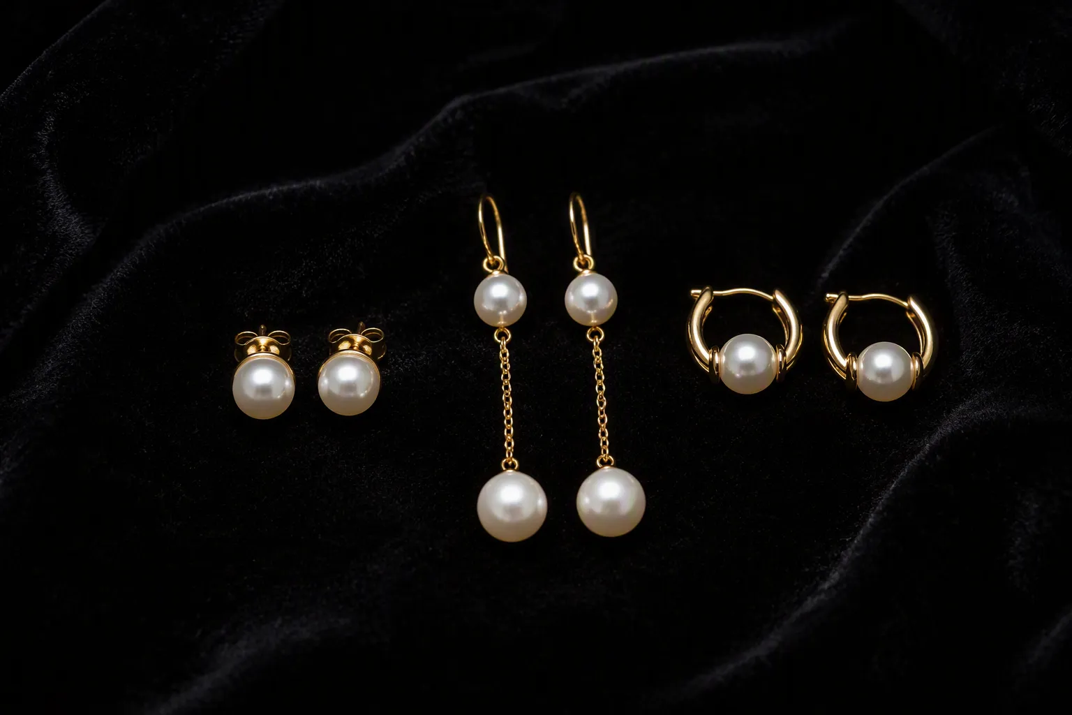pendientes oro perlas - Fotografía editorial de tres pares de pendientes oro perlas sobre terciopelo negro: un par de studs clásicos, unos colgantes largos y unos aros con perla central, con etiquetas de material y acabado