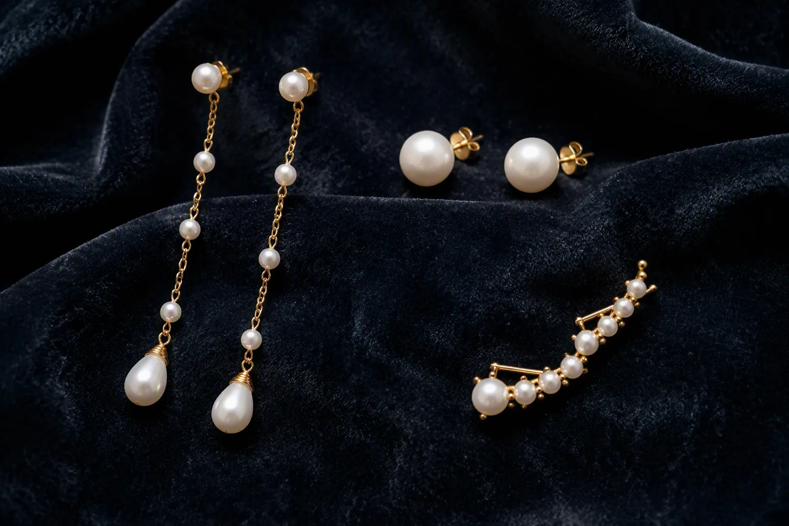 pendientes oro y perlas - Fotografía editorial de tres estilos de pendientes oro y perlas dispuestos sobre terciopelo oscuro: un modelo colgante largo, unos studs clásicos y un diseño trepador, para ilustrar la variedad de formatos disponibles