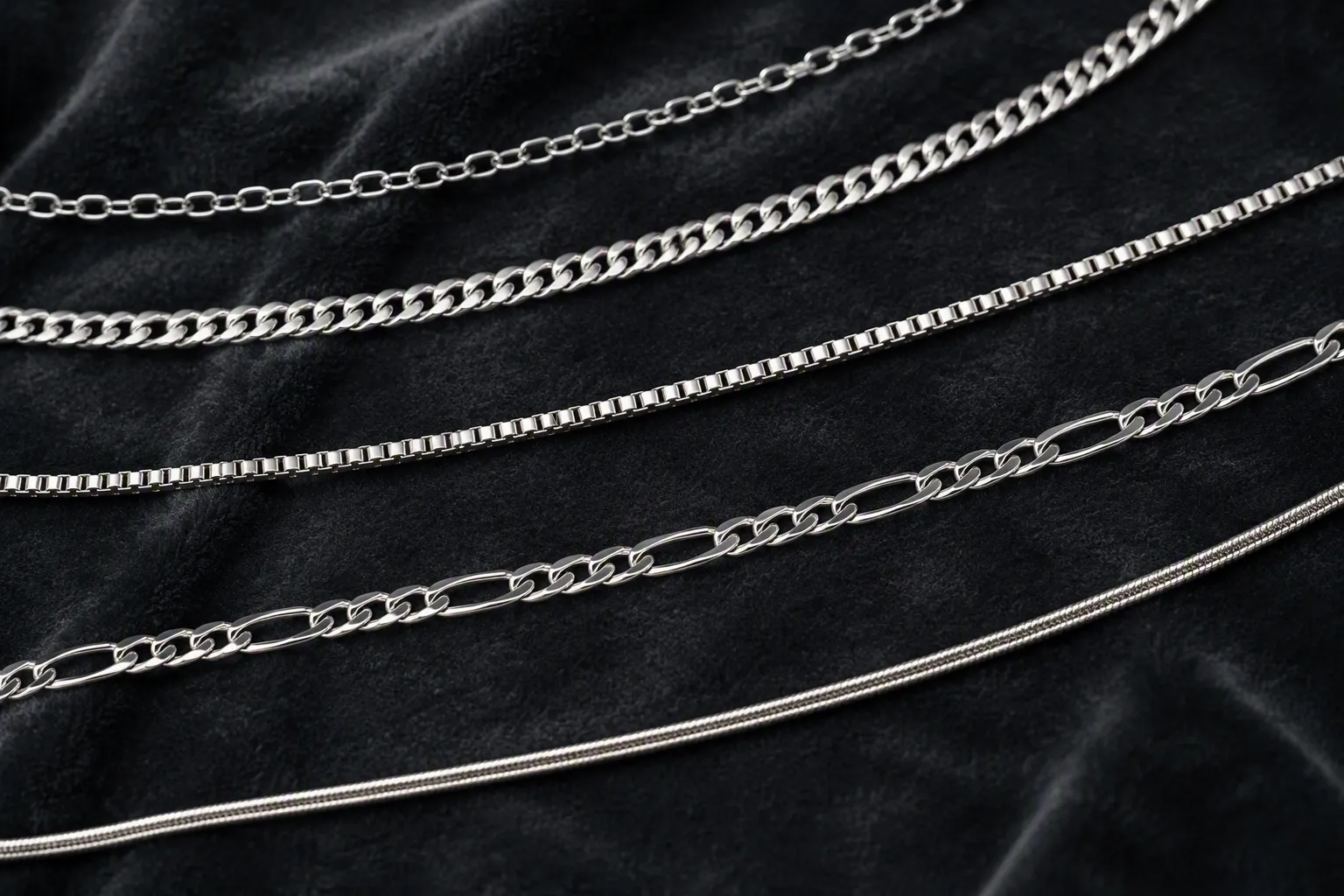 sterling silver necklace wholesale - Photorealistic flat-lay photograph of five .925 sterling silver necklaces arranged on a dark charcoal velvet surface — cable chain, curb chain, box chain, figaro, and snake chain styles each clearly distinguishable, soft overhead studio lighting casting gentle shadows that reveal individual link texture and silver sheen, no text or labels, documentary product photography feel