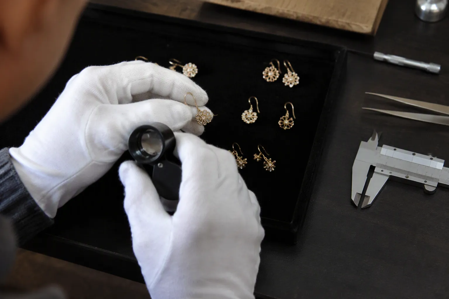 bisutería joyería - Imagen de proceso de control de calidad en taller de bisutería: manos con guantes inspeccionando pendientes sobre bandeja de fondo negro, con lupas y calibres visibles