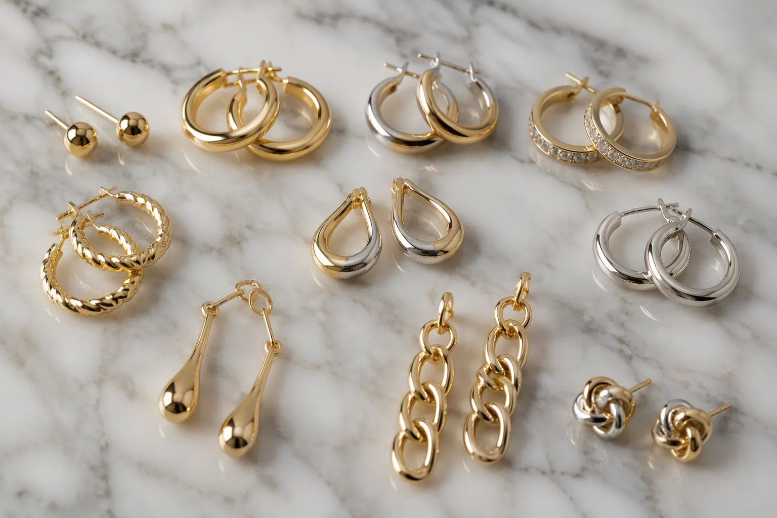 pendientes bañados en oro - Primer plano de varios pendientes bañados en oro sobre superficie de mármol blanco — studs, argollas y colgantes — mostrando variación de estilos y acabados en colores dorado, plata y bicolor