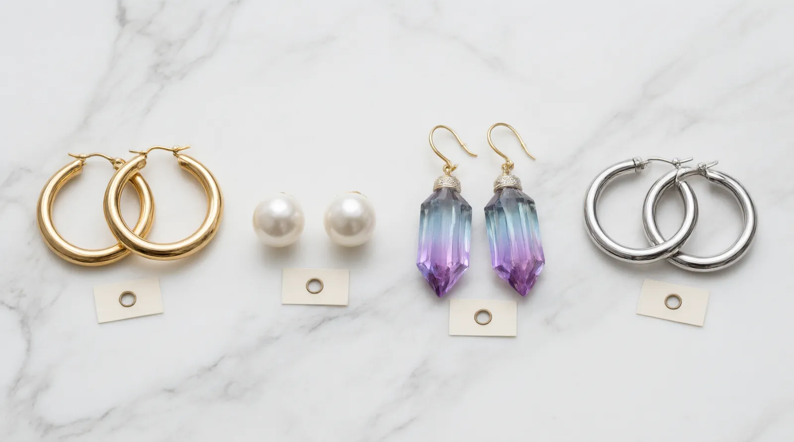 pendientes bisutería - Fotografía editorial comparativa sobre mármol blanco de cuatro tipos de pendientes bisutería mujer: aros dorados lisos, studs de perla sintética, pendientes gota con cristal degradado y set de aros plateados tipo plata, con pequeñas etiquetas de material visibles junto a cada pieza
