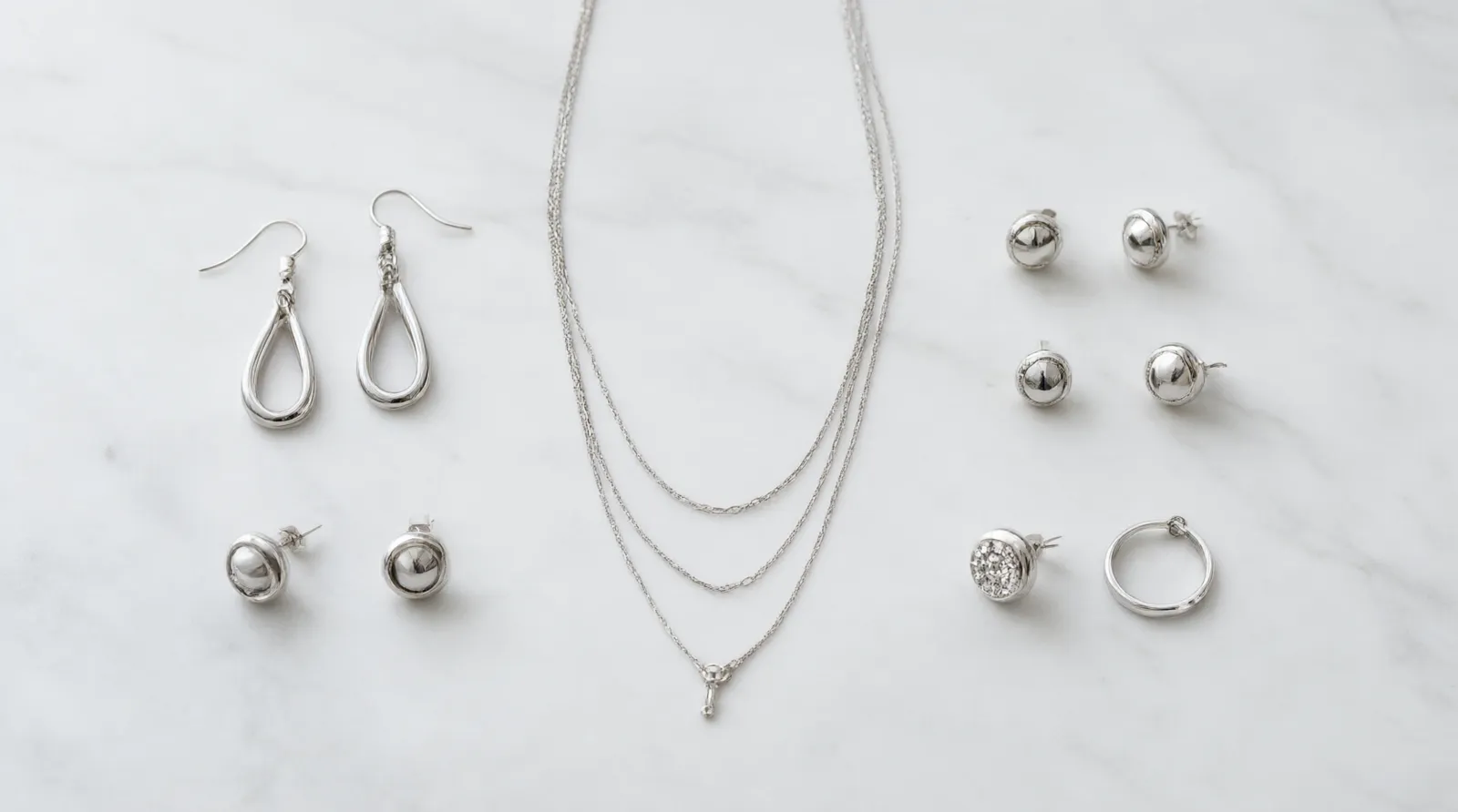 silberner schmuck - Photorealistic flat-lay product shot of a curated silver-toned jewelry assortment – rhodium-plated drop earrings, stud earrings, a delicate layered necklace and a rhinestone ring – arranged symmetrically on a matte white marble surface, soft diffused daylight from above, clean documentary product photography with shallow depth of field