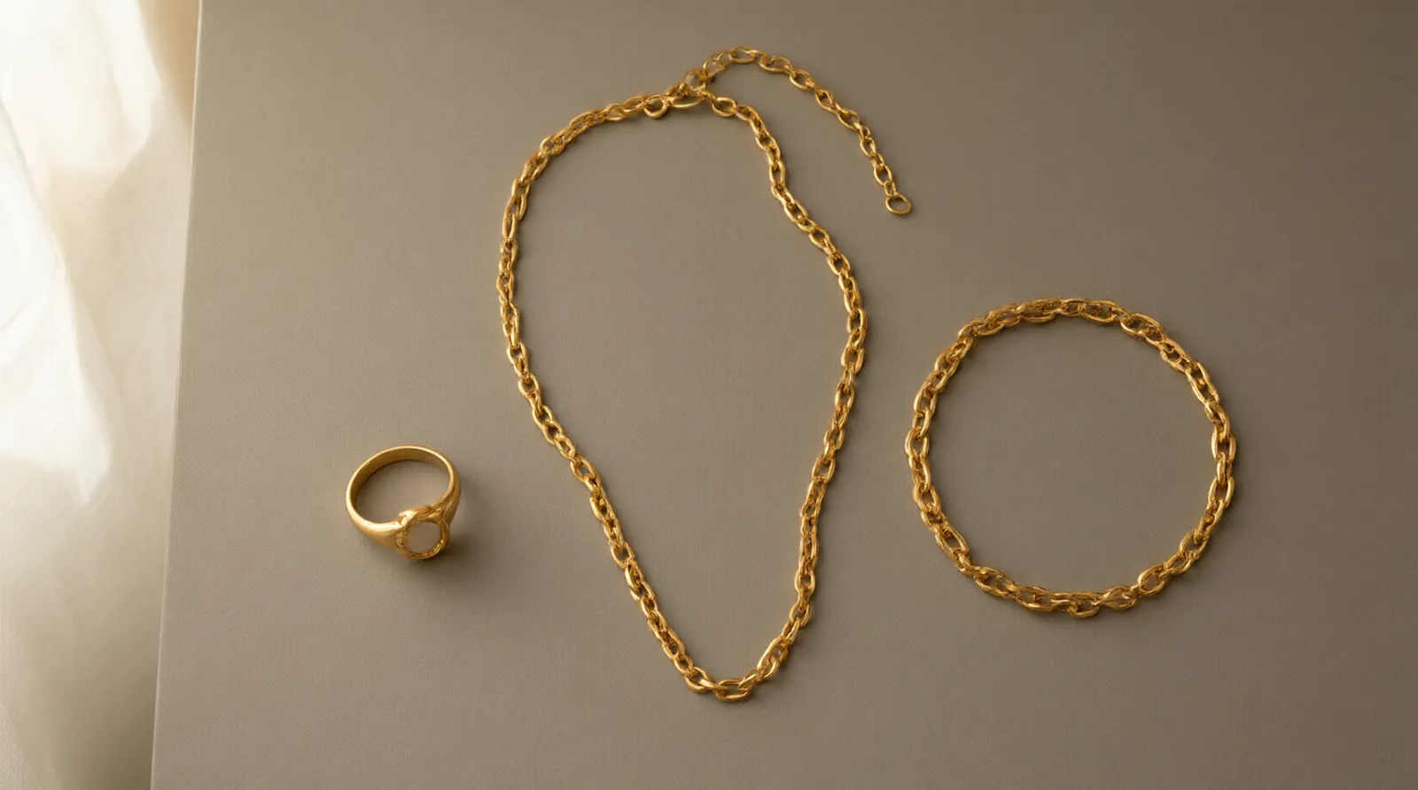 14k gold pvd - Photorealistic flat-lay of three jewelry pieces on a matte warm-gray surface — a chain necklace, a ring, and a bracelet, all in a warm gold tone — arranged in a triangular composition, soft diffused natural light from a window to the left, authentic product presentation style, no text or overlays