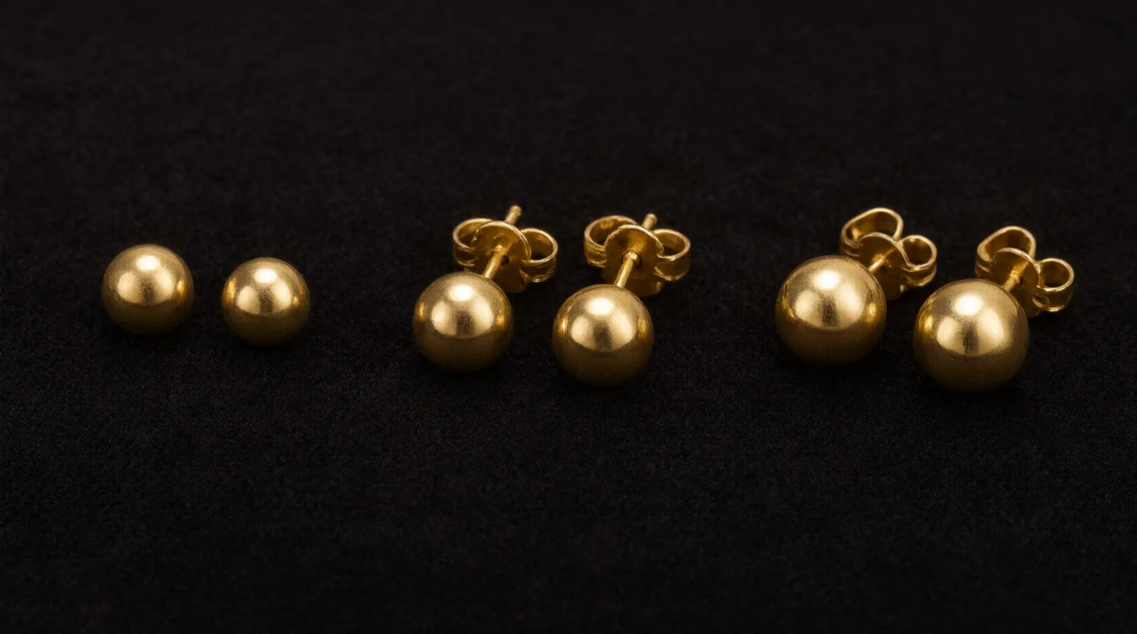 pendientes bola oro - Fotografía editorial macro que muestra tres pares de pendientes bola oro de diferentes diámetros (3 mm, 5 mm y 7 mm) alineados sobre una bandeja de terciopelo oscuro, destacando el brillo del acabado liso y el mecanismo de cierre de presión visible en el par central