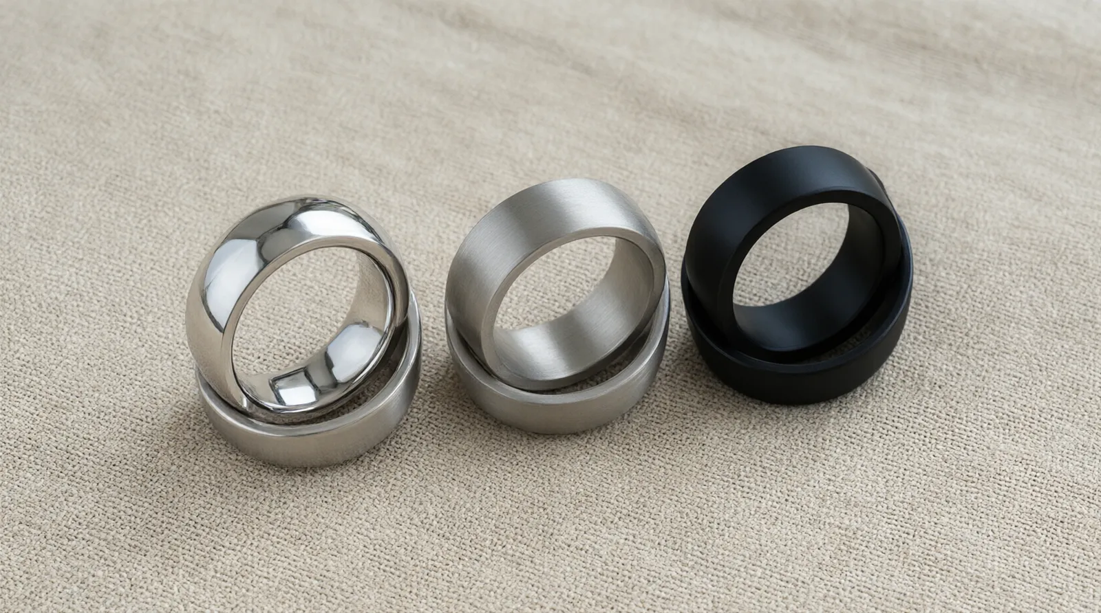 stainless steel ring - Editorial conceptual photo showing three different stainless steel ring finishes—a mirror polish, a brushed matte, and a black PVD coating—arranged in a row on a piece of raw linen fabric, highlighting the textural contrast, natural diffused daylight