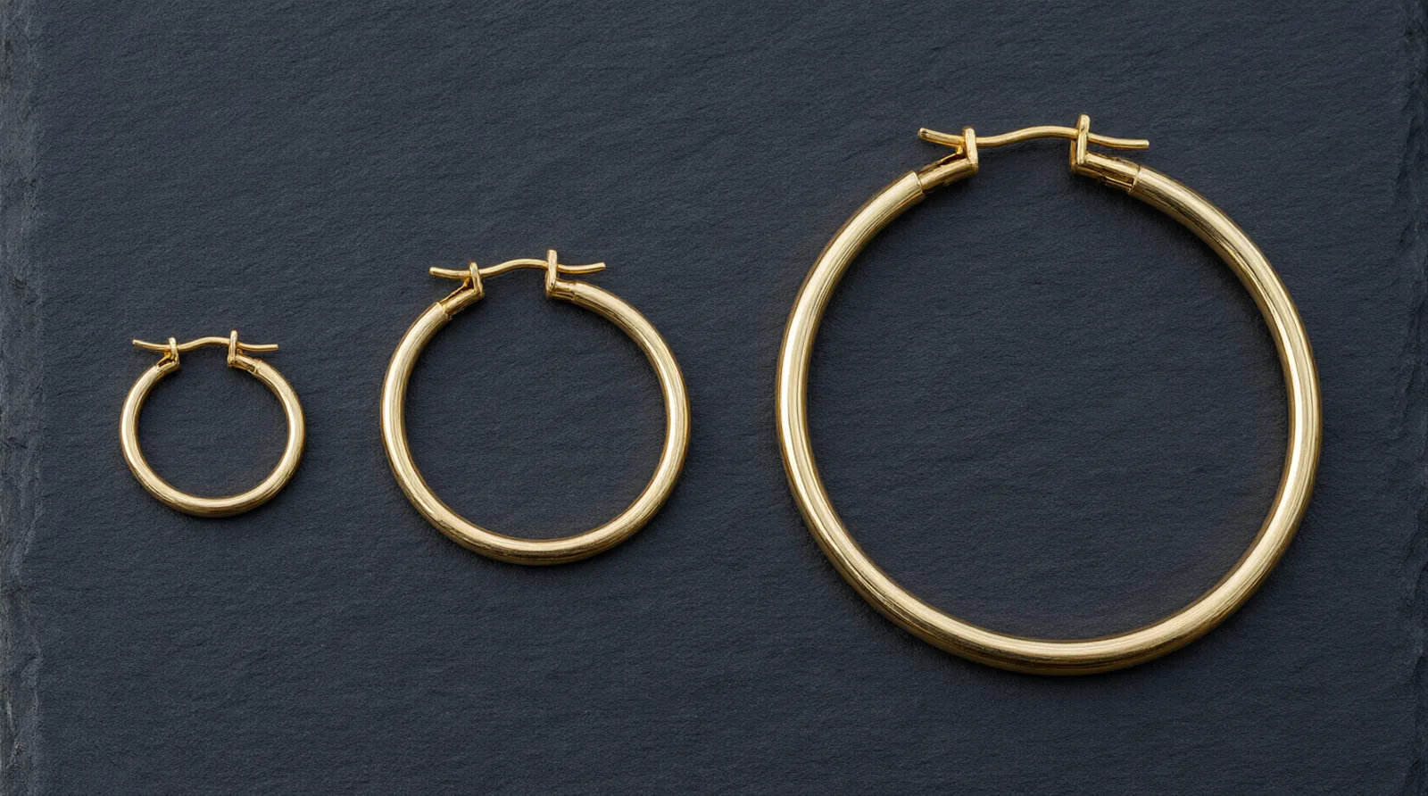 14k gold hoop earrings - A photorealistic macro shot showing three 14k gold hoop earrings of varying diameters (15mm, 30mm, 50mm) laid flat on a dark slate surface, highlighting the difference in hinge mechanisms and wire thickness